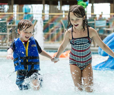 A young brother and sister are holding hands laughing as they run around and play in the water.