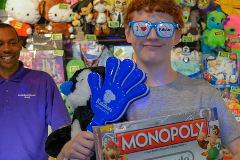 A kid showing his prizes, a monopoly board game, sweet sunglasses and large hand clappers.