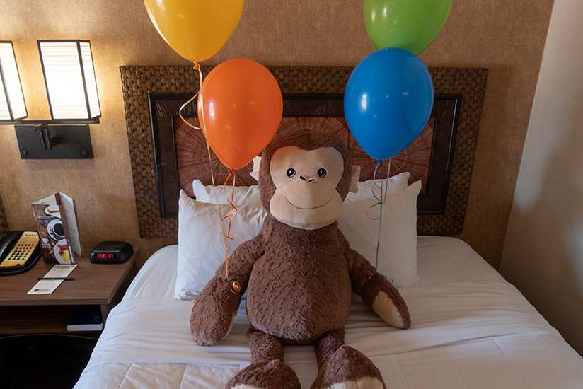 Jumbo Monkey Plush sitting on a bed in a Kalahari Guest Room with four Colorful Balloons