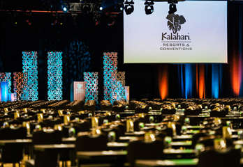 Inside Kalahari's convention center, rows of tables and chairs face a large screen hanging from the ceiling with sculptural lighting behind a podium.