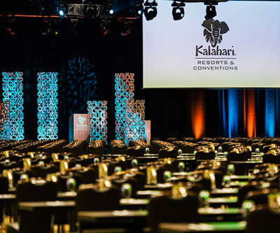 Inside Kalahari's convention center, rows of tables and chairs face a large screen hanging from the ceiling with sculptural lighting behind a podium.